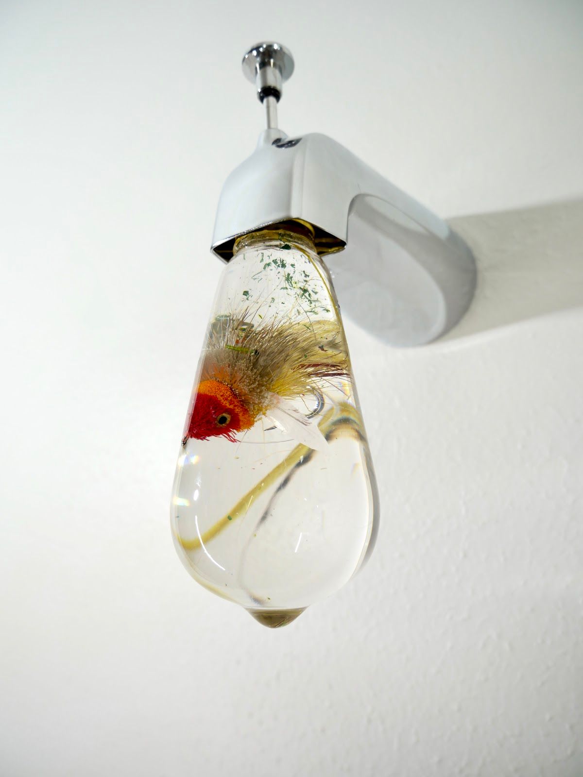 Chrome faucet seen from below, its resin bulb holding a red-and-orange fly with a bead eye