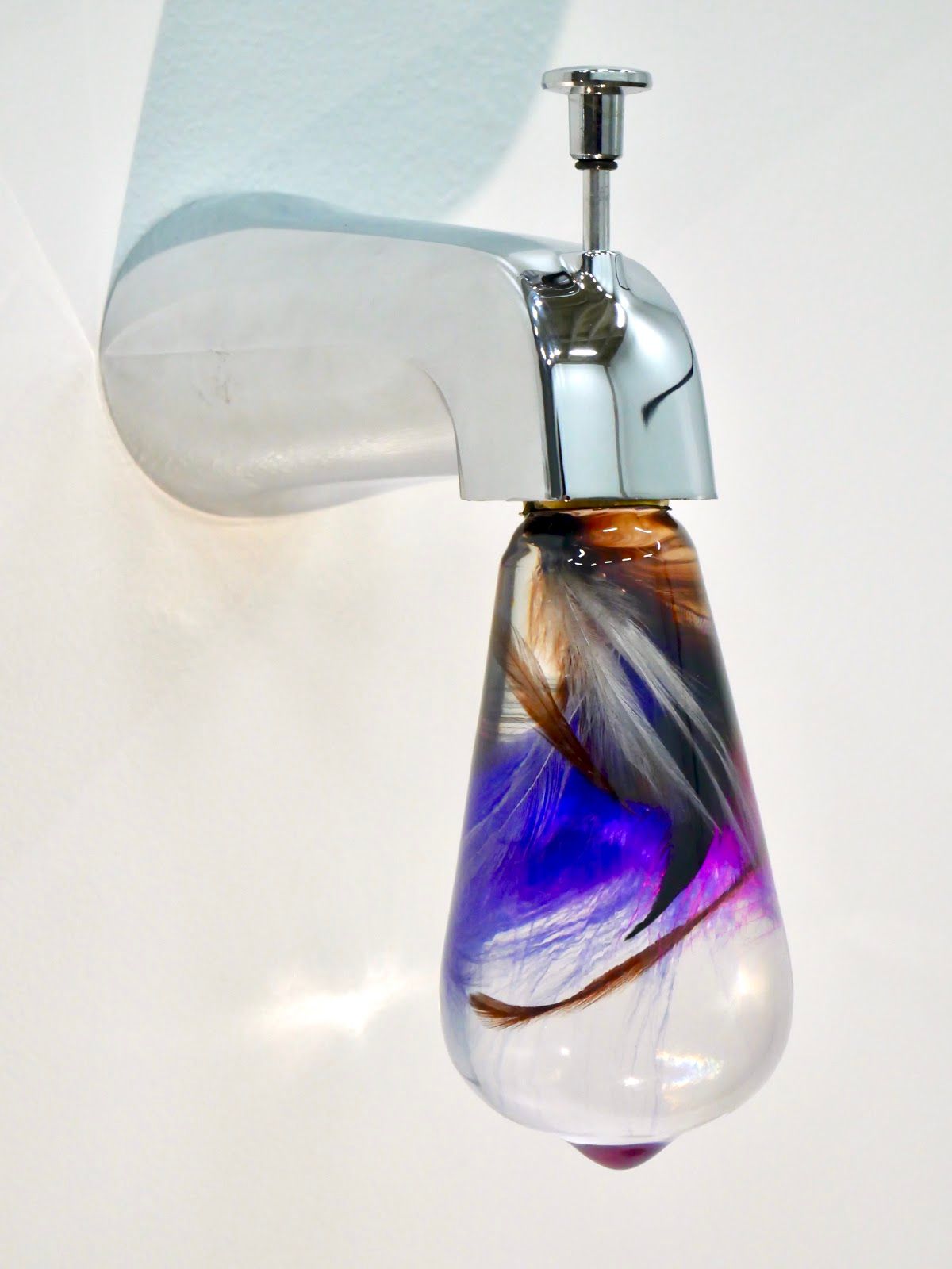 Close-up of a chrome faucet ending in a clear resin bulb with violet, magenta and brown feathers