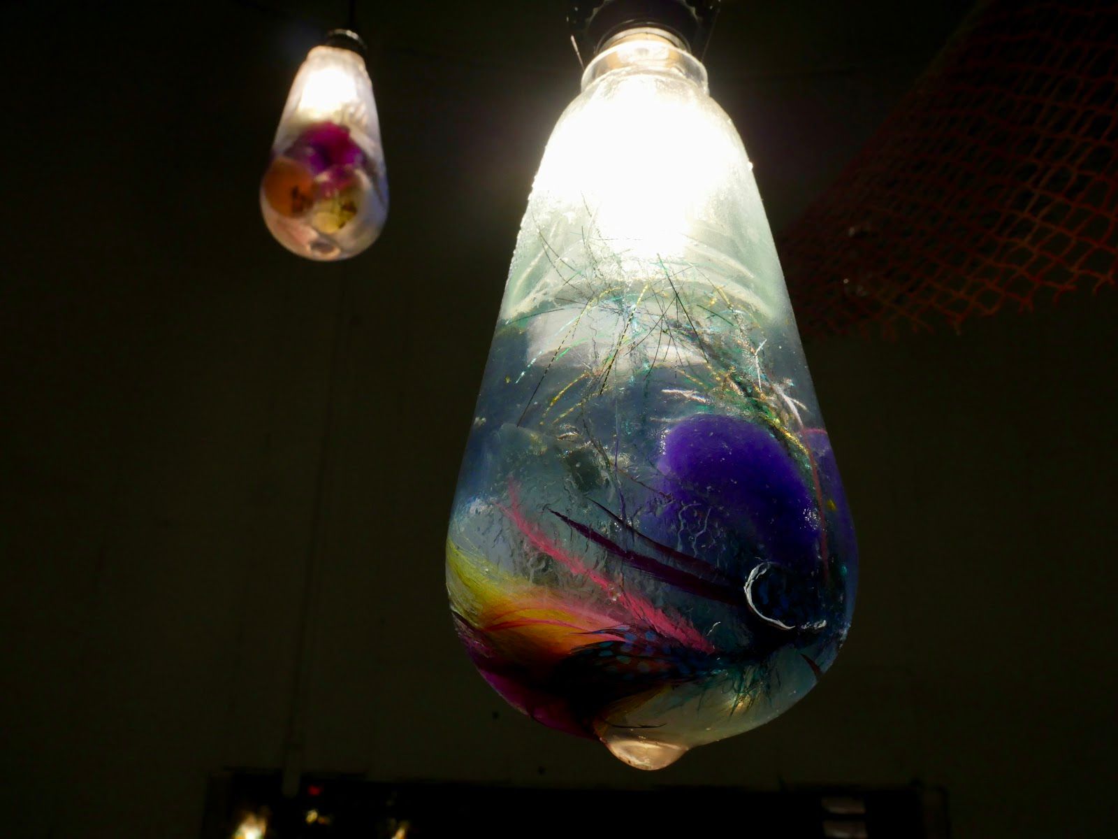 Close view of two glowing resin pendant bulbs with swirling violet and magenta fly feathers suspended inside