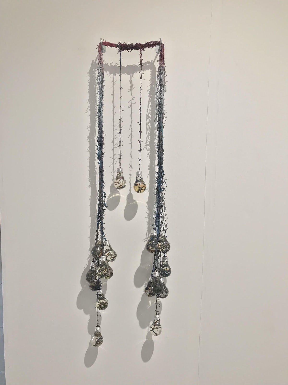 Wall-hung sculpture of spray-painted fishnet strands descending from a horizontal bar, ending in clusters of small resin light bulbs