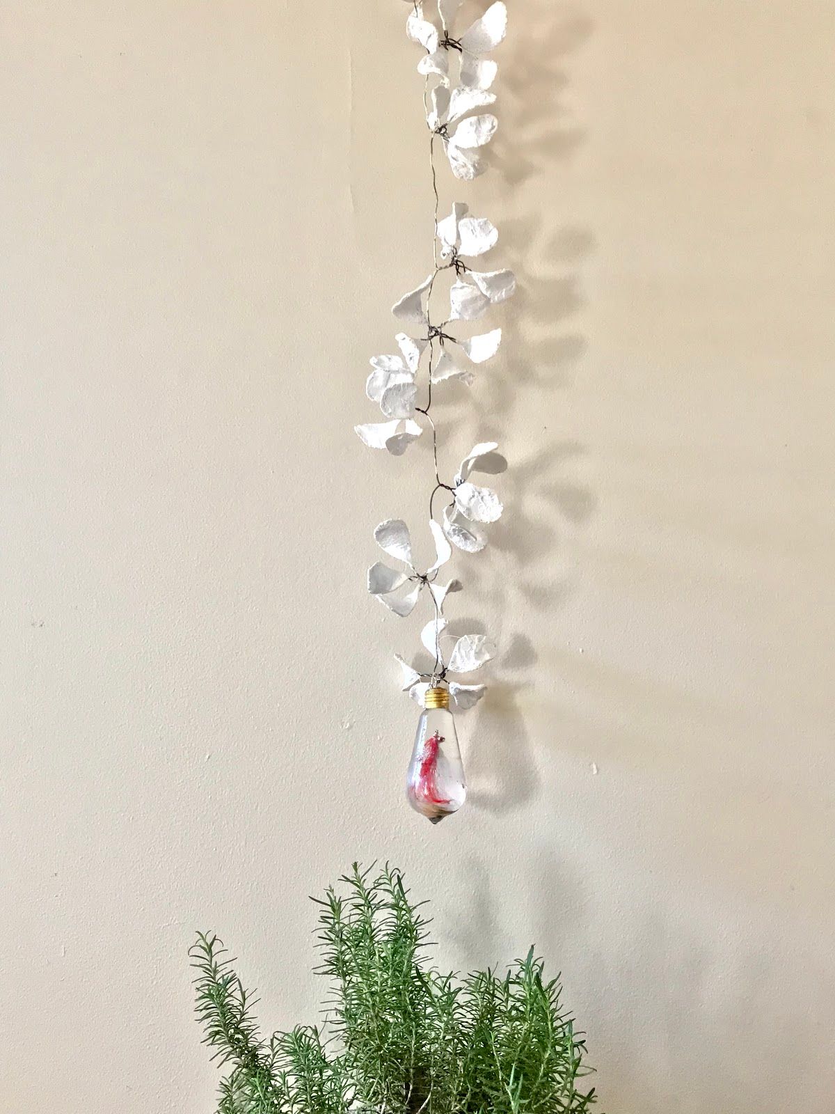A vertical vine of white plaster flowers on wire hanging against a cream wall, ending in a small clear resin bulb with a red fly