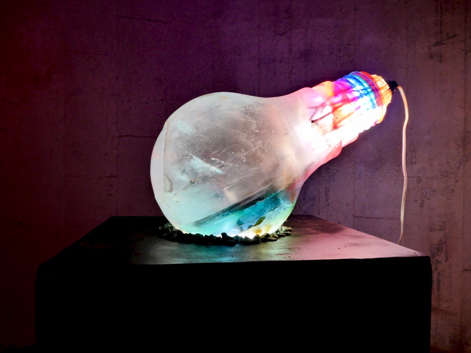 The same oversized resin bulb on a black plinth against a purple concrete wall, lit from within in aqua and rainbow tones