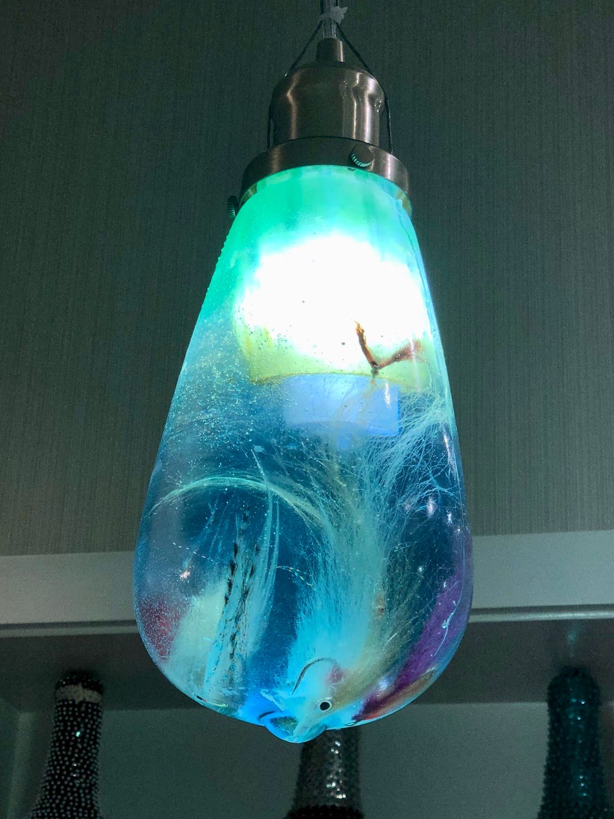 Glowing turquoise resin pendant bulb hanging from a brushed-steel fitting, white feathered fishing flies swirling inside