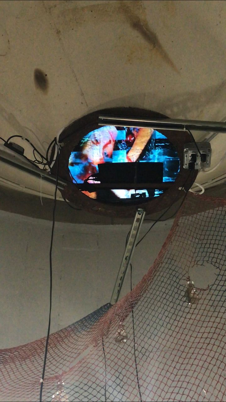 Circular opening in a concrete silo ceiling showing a fragmented blue and red video, with red netting draped below