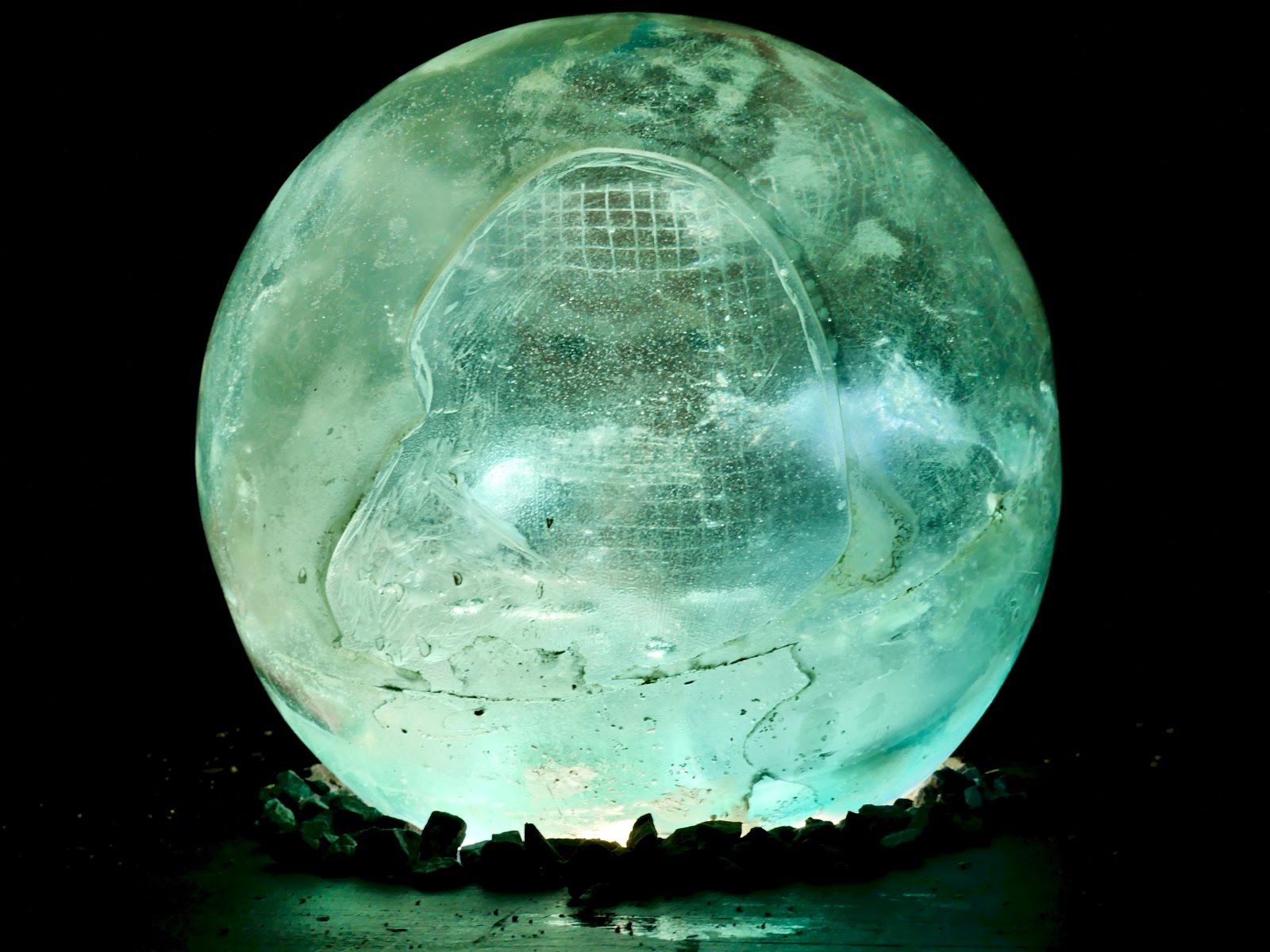 The glowing resin sphere photographed in darkness, luminous sea-green, resting on dark stones