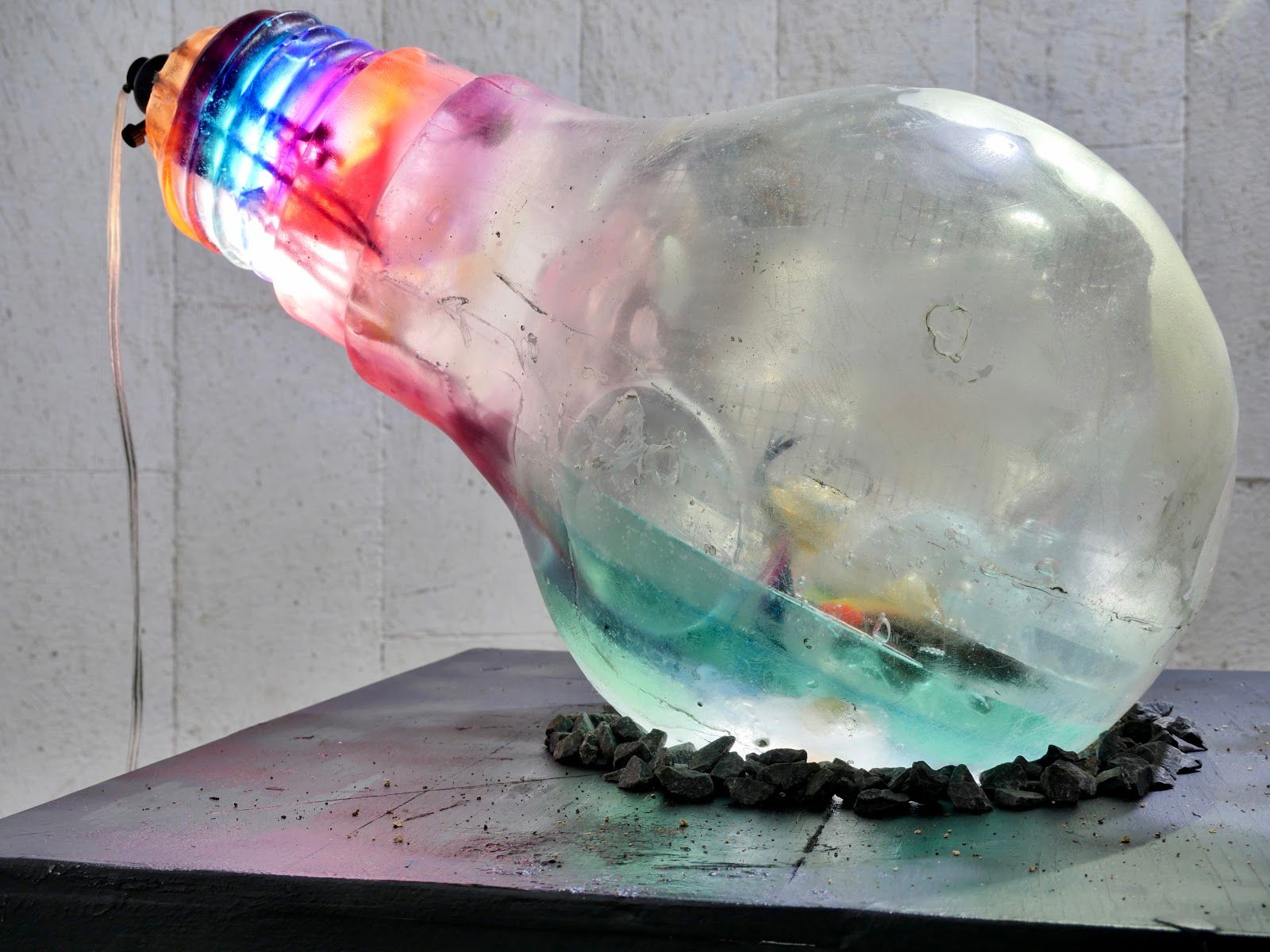 Large clear resin light bulb lying on its side on a dark pedestal, its neck glowing in bands of pink, blue and orange, resting on a ring of dark stones
