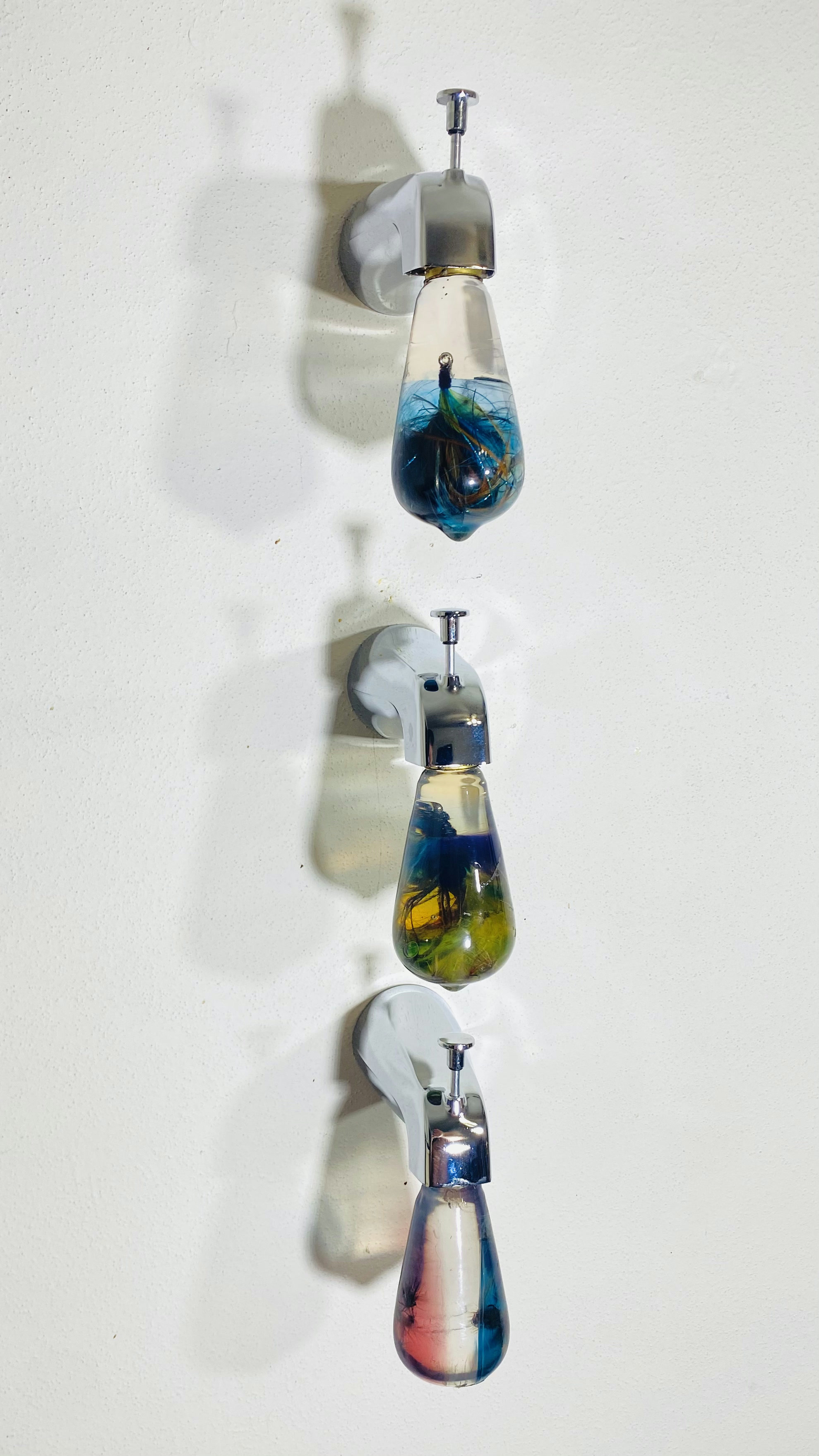 Three chrome wall faucets stacked vertically, each ending in a teardrop resin bulb filled with water and colorful fishing flies