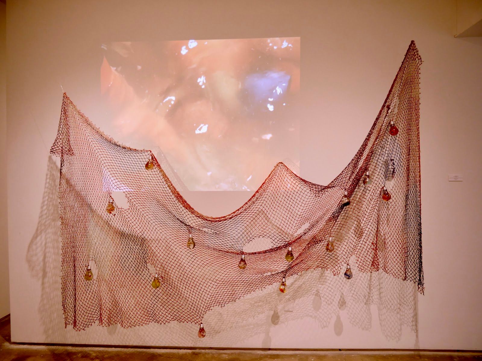 Rust-red fishing net draped in a wave across a gallery wall, dotted with small amber and red resin light bulbs, a pink underwater video projected behind it